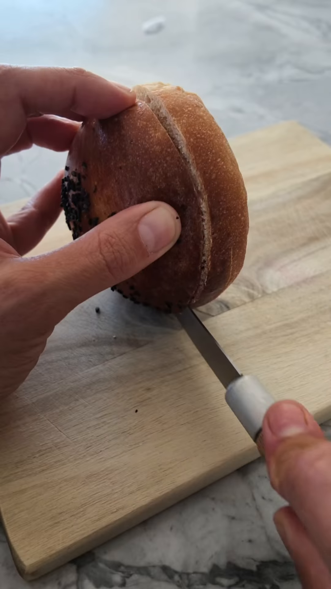 Bun à burger au levain doré parsemé de graines de sésame noir, tranché en deux au couteau sur une planche en bois, révélant une mie fine et serrée
