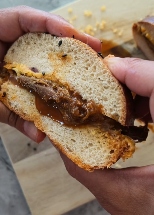 Burger maison ouvert dans les mains, échine de porc confite nappée de sauce, cheddar fondu et buns au levain dorés saupoudrés de graines de sésame
