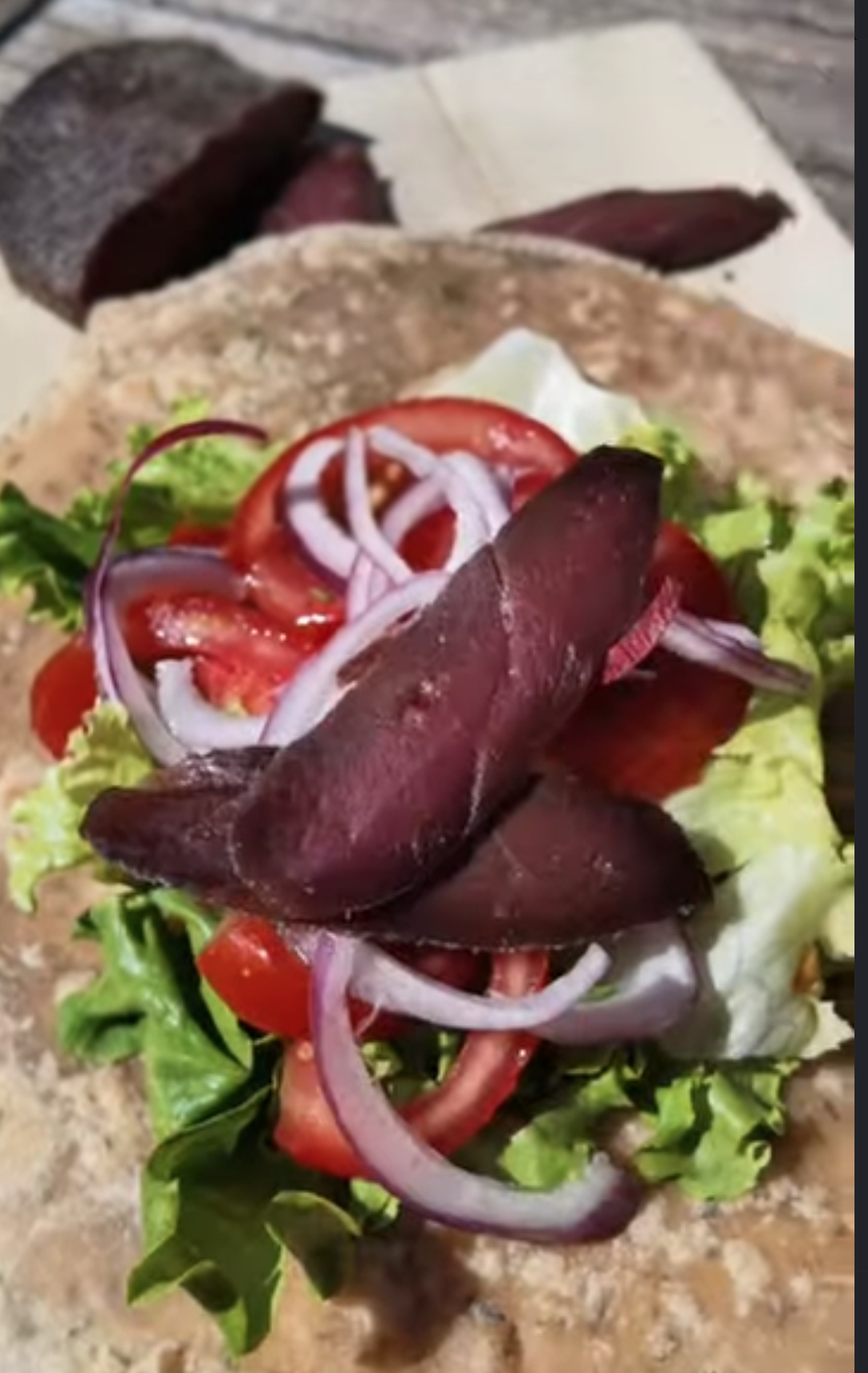 Fines tranches de gravelax de magret de canard rouge sombre déposées sur une salade de tomates, oignons rouges et jeunes pousses dans un wrap
